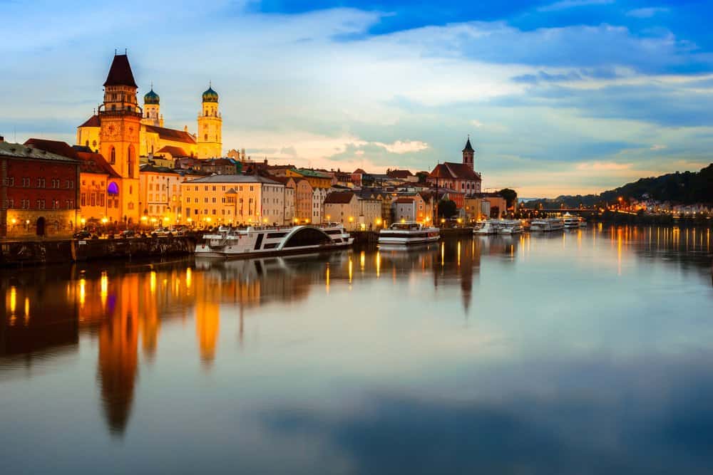 Scenic evening view of Passau, Germany, with historic architecture and St. Stephen’s Cathedral reflecting on the Danube River, illuminated by golden lights.