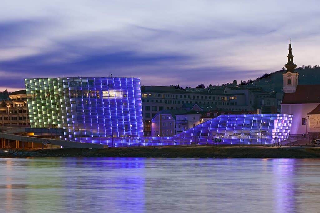 The Ars Electronica Center in Linz, Austria, illuminated in vibrant blue lights at night, reflecting on the Danube River. A hub for digital arts and technology.