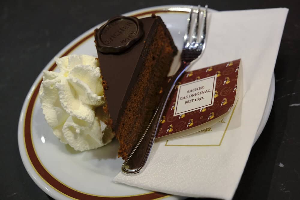 A slice of the Original Sacher-Torte from Vienna, Austria, served with whipped cream on a classic plate, featuring its signature chocolate glaze and official seal.