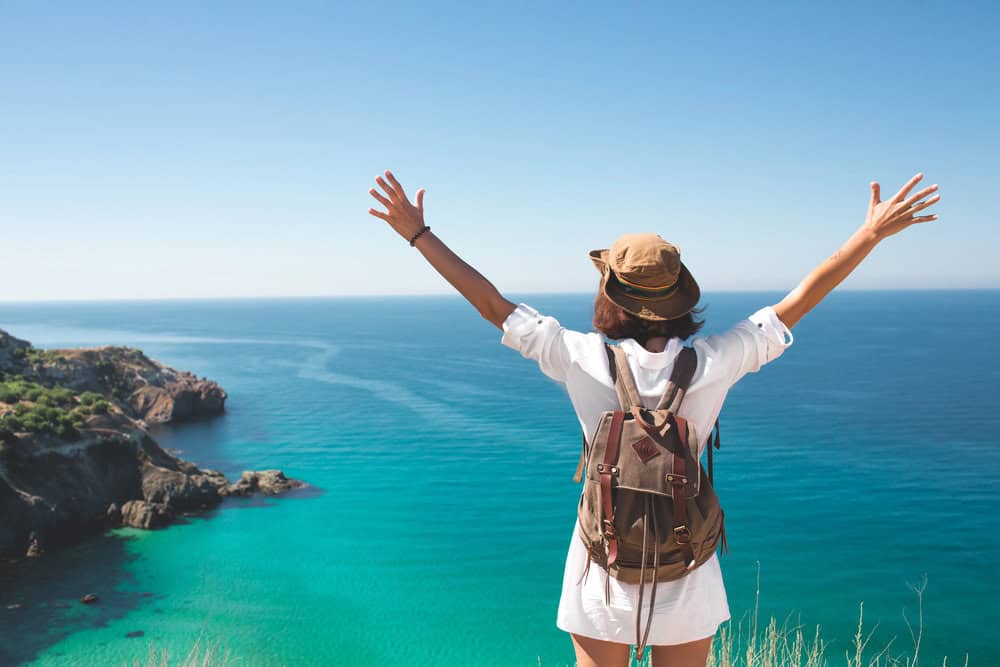 A traveler wearing a backpack stands on a scenic cliffside overlooking a turquoise ocean, arms raised in freedom. This image captures the joy and ease of packing light for travel.