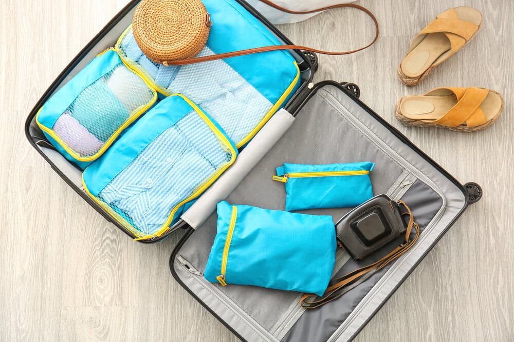 An open suitcase neatly packed with blue packing cubes, showcasing an organized way to pack light for travel. Nearby are a woven handbag, a compact camera, and comfortable sandals, reinforcing the benefits of efficient packing.