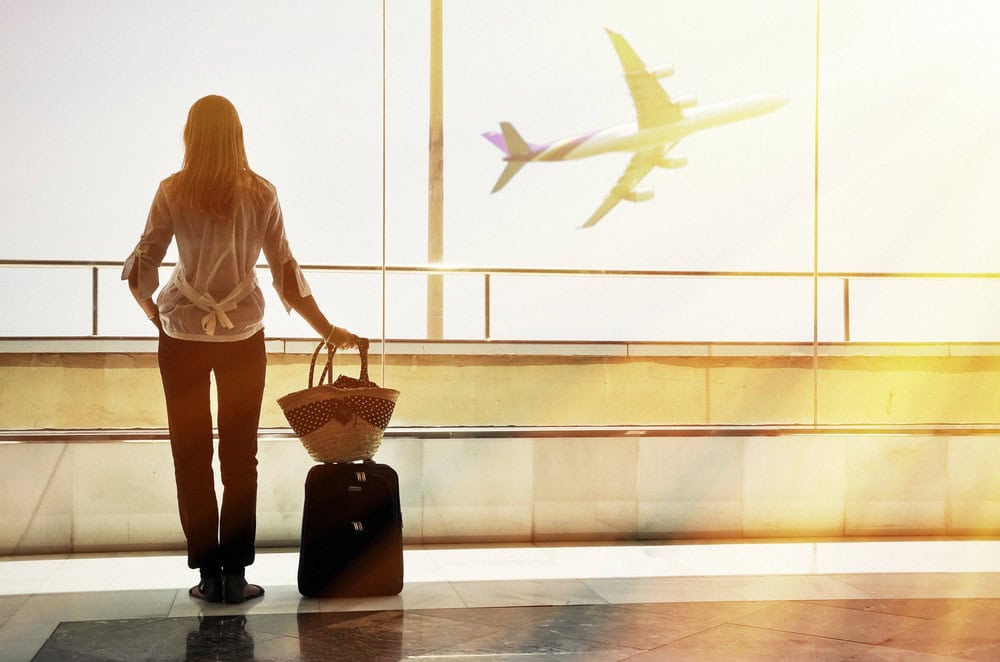 A woman standing at an airport window with a small carry-on suitcase and a woven travel bag, watching an airplane take off. The sunlight streams through the glass, highlighting the ease and freedom of packing light for travel.