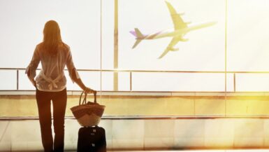 A woman standing at an airport window with a small carry-on suitcase and a woven travel bag, watching an airplane take off. The sunlight streams through the glass, highlighting the ease and freedom of packing light for travel.