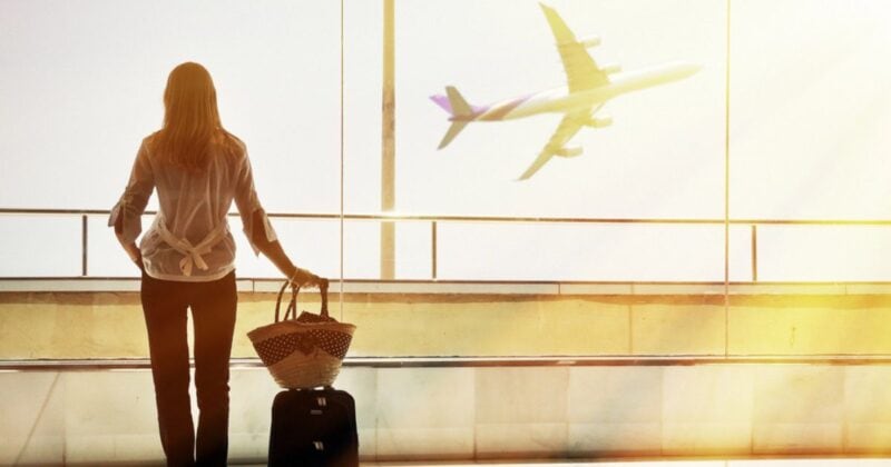 A woman standing at an airport window with a small carry-on suitcase and a woven travel bag, watching an airplane take off. The sunlight streams through the glass, highlighting the ease and freedom of packing light for travel.